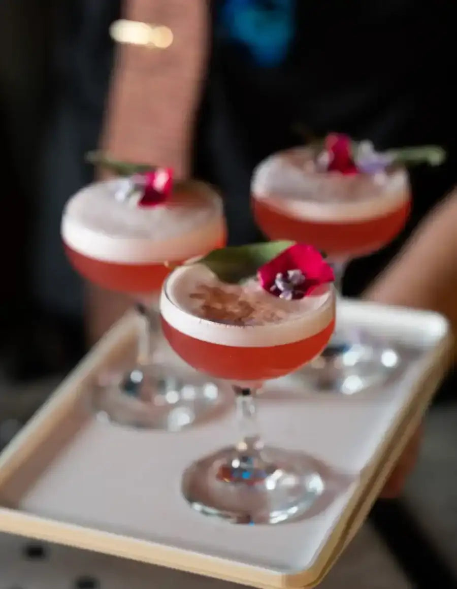 Three elegant coupe glasses filled with a vibrant red cocktail, each topped with a delicate white foam layer and garnished with a fresh flower.