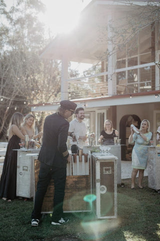 Outdoor cocktail class with Trolley'd bar setup, as a bartender serves guests in a sunlit garden party, creating a lively and elegant atmosphere.