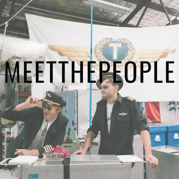 Two men dressed in black pilot uniforms with gold epaulets and winged insignia stand behind a counter under a banner that reads ’meet the people.’