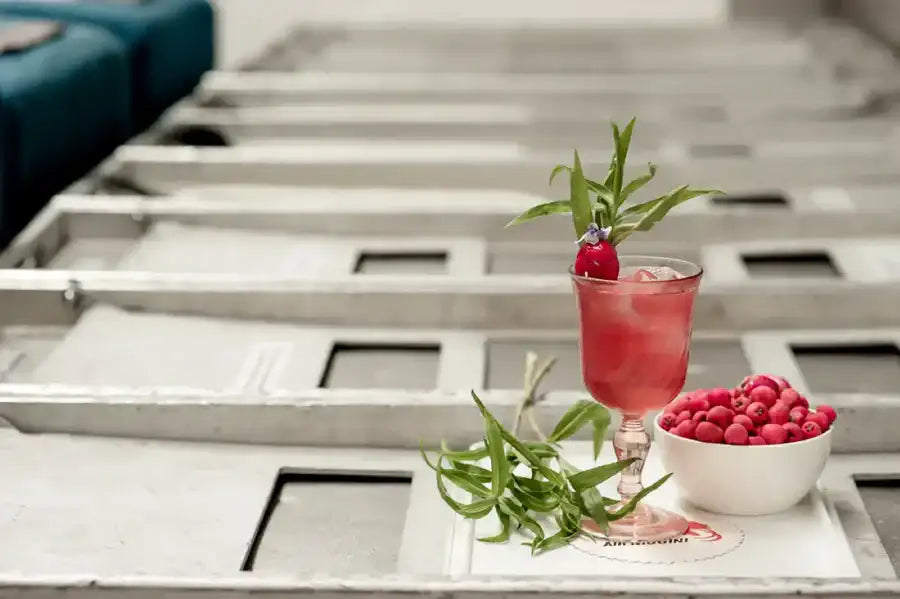 A vibrant red cocktail in a stemmed glass, garnished with a raspberry and fresh green herbs.
